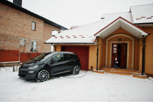 An electric vehicle parked outside a snow-covered home, charging via a cable connected to the house, with a child standing at the entrance.