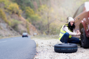 Woman crouching next to her car, inspecting a tire with a concerned expression on a quiet road.