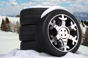 Stack of winter tires and wheels covered with snow, placed in a winter landscape with mountains in the background.