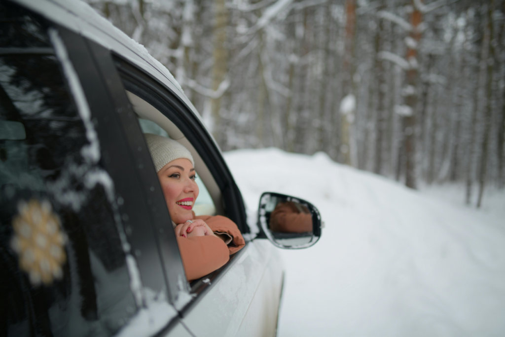 Person leaning out of a car window, smiling while surrounded by a snowy forest.