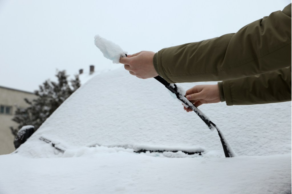 Close-up of someone clearing snow and ice from a car’s windshield wiper on a snowy winter day.